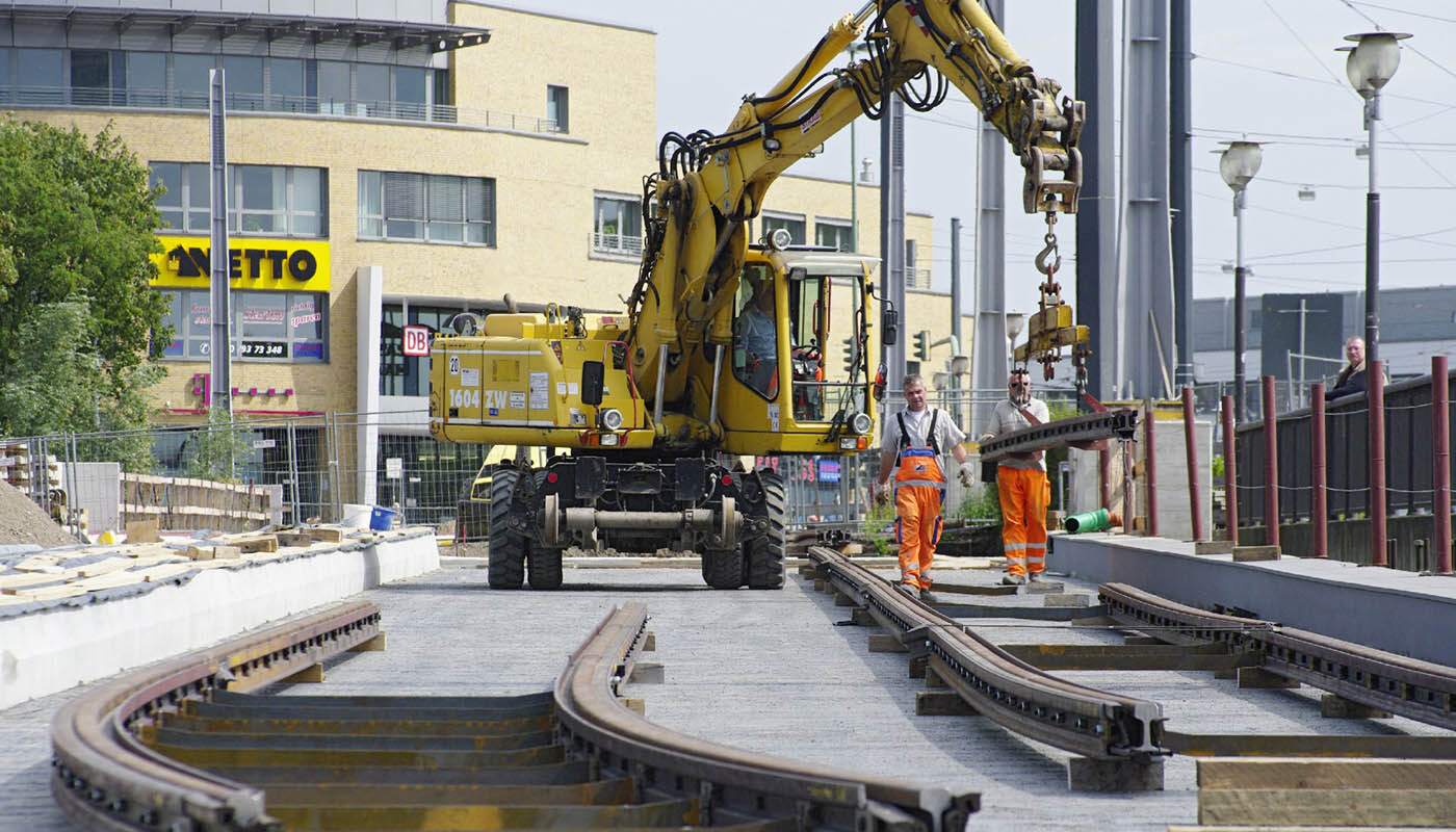 Foto: Drei Männer bei den Gleisbauarbeiten, der Arm eines Baggers bewegt die schweren Gleiselemente, im Hintergrund das Bahnhofsgebäude.