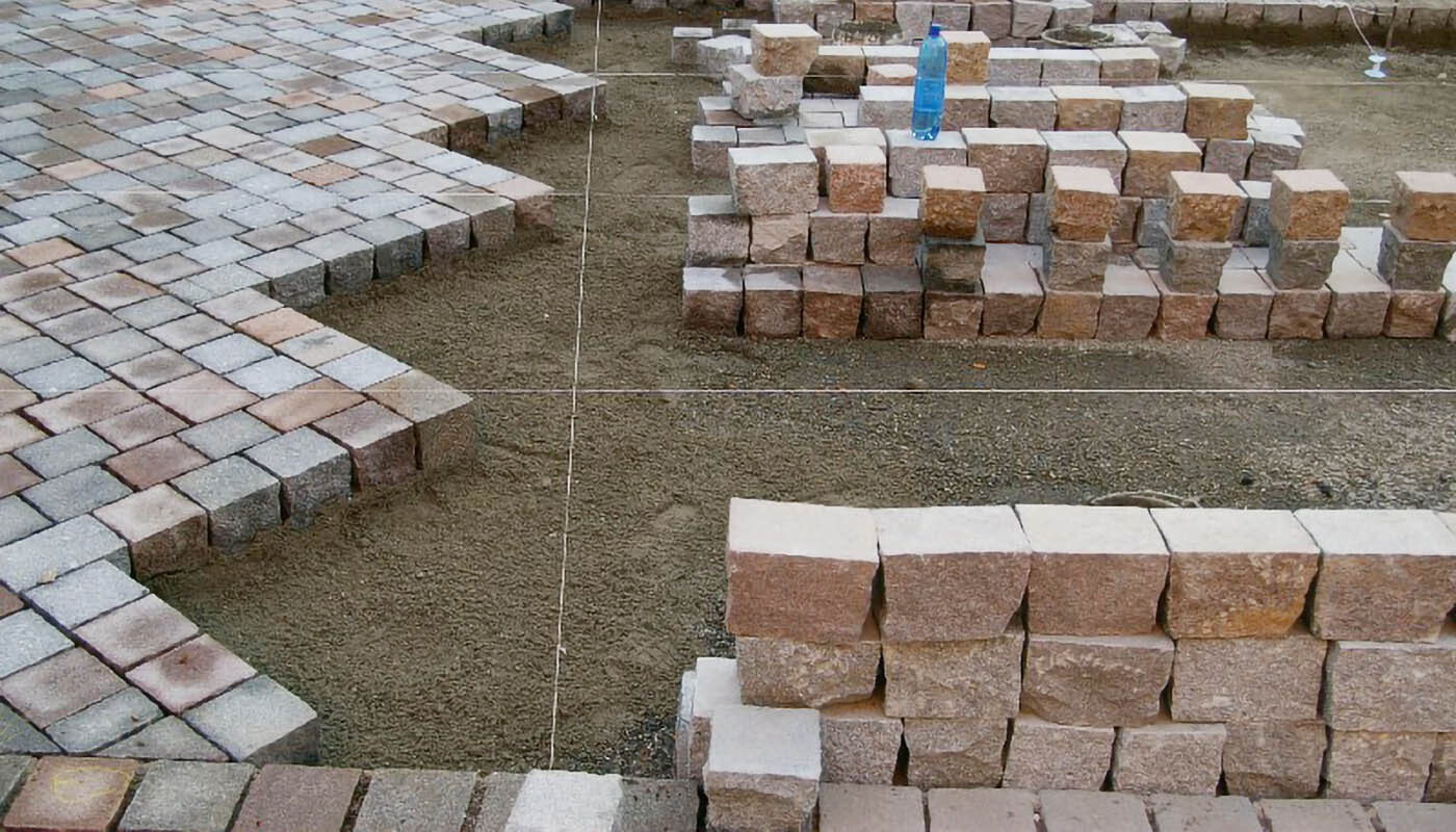 Foto: Quadratische Pflastersteine, die einen Diagonalverband bilden, aufgespannte Schnüre und bereitgelegte Pflastersteine.