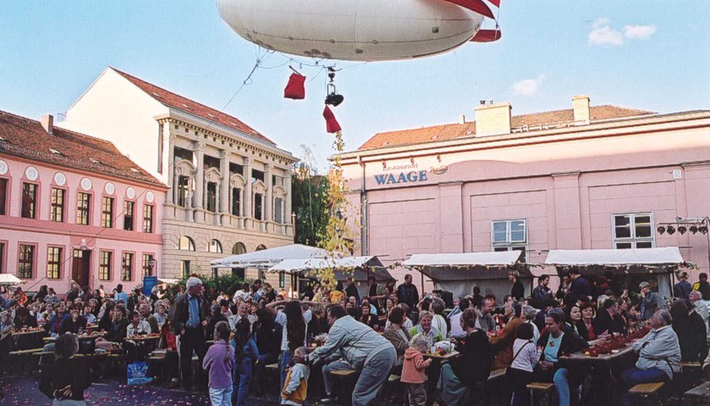 Foro: Der belebte Neue Markt mit Marktständen, besetzten Tischen und Bänken. Über dem Geschehen ein großer Ballon in Form eines Zeppelins.
